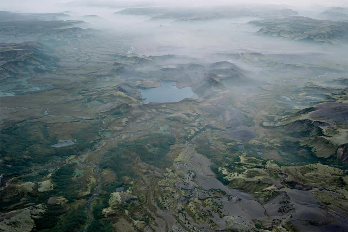 A broad volcanic basin, seen from above, its low valleys and crater-lake terrain softened by faint layers of haze. Quiet overcast daylight reveals realistic topographic detail in muted deep greens, slate blues, and ash gray. Wide aerial composition, restrained documentary illustration, subtle archival grain, and no dramatic effects.
