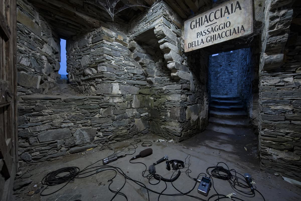 An empty stone chamber beside the old icehouse passage, microphones and cables arranged quietly on the floor. Restrained documentary illustration of Castello di Montebello at dusk, framed in a single wide view with muted slate blue, weathered stone and charcoal, low atmospheric light, subtle archival grain, realistic and non-sensational.