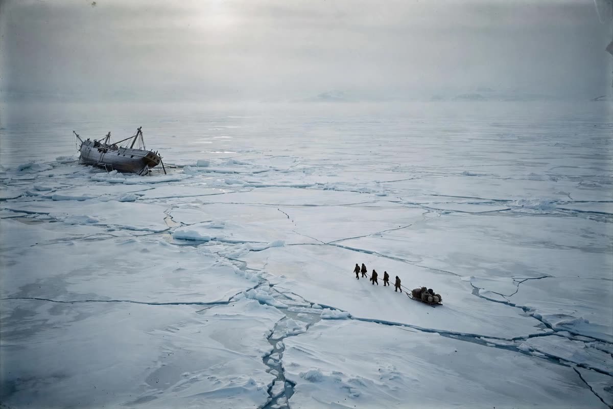 Distant men leave a listing wooden ship, dragging salvaged equipment across a cracked ice floe, their figures tiny against the vast Antarctic expanse. Low polar daylight filters through fine haze over wind-scoured snow. Wide, distant composition with dramatic negative space; cinematic historical illustration with restrained documentary realism, cold glacial blues, slate grays, muted ivory, and subtle warm accents, rendered with fine-grain film texture.